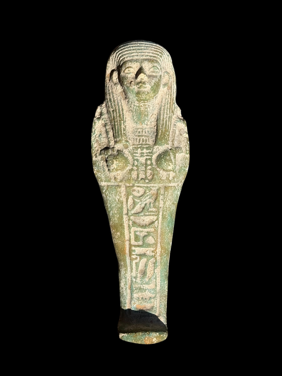 Front view of a green and pale mottled shabti figurine with a striped wig, crossed arms and a vertical panel of hieroglyphic details.