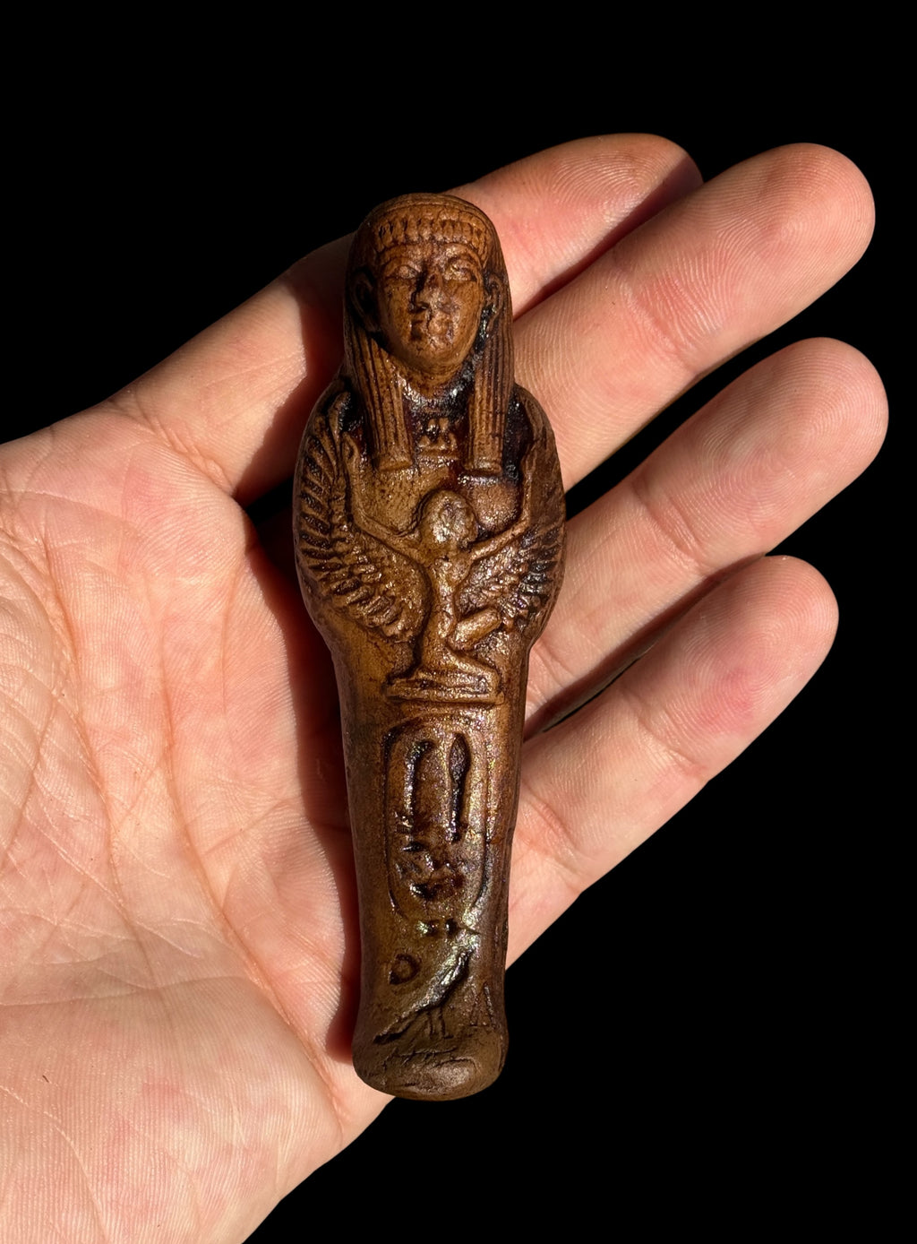 Brown Egyptian stone figurine with winged Isis resting in an open palm, showing the face, striped headdress, winged Isis motif and markings on the front.
