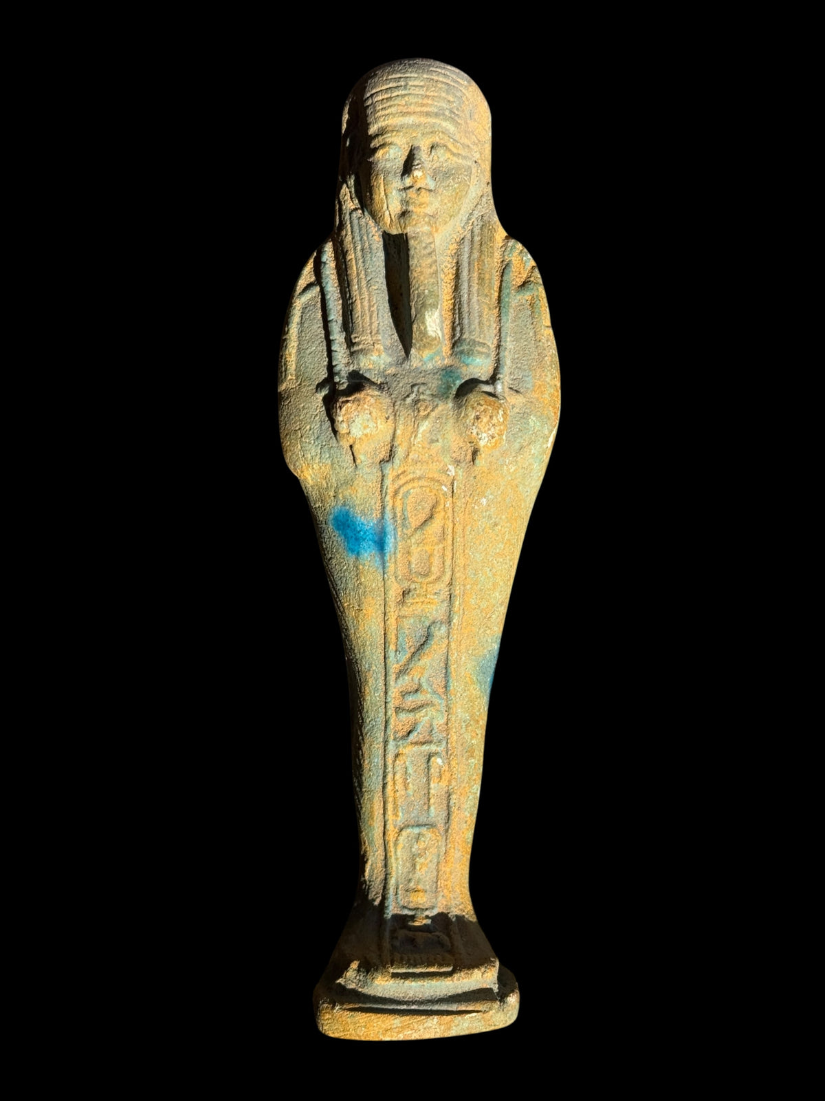 Front view of a shabti figurine with a striped headdress, long beard, incised front panel and ochre and blue-green surface.