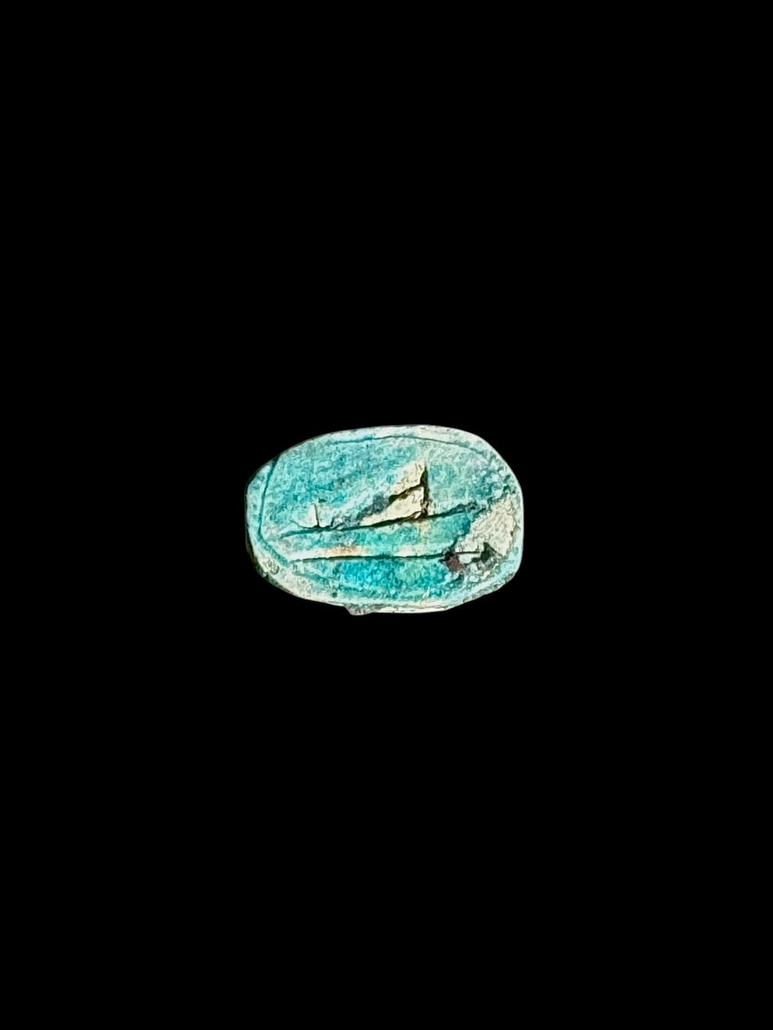 Underside of a turquoise-coloured stone scarab with incised markings, pale patches and an oval border against a black background.