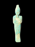 Front view of a pale turquoise and cream shabti with a tall crown, straight beard, crossed arms and incised markings against a black background.