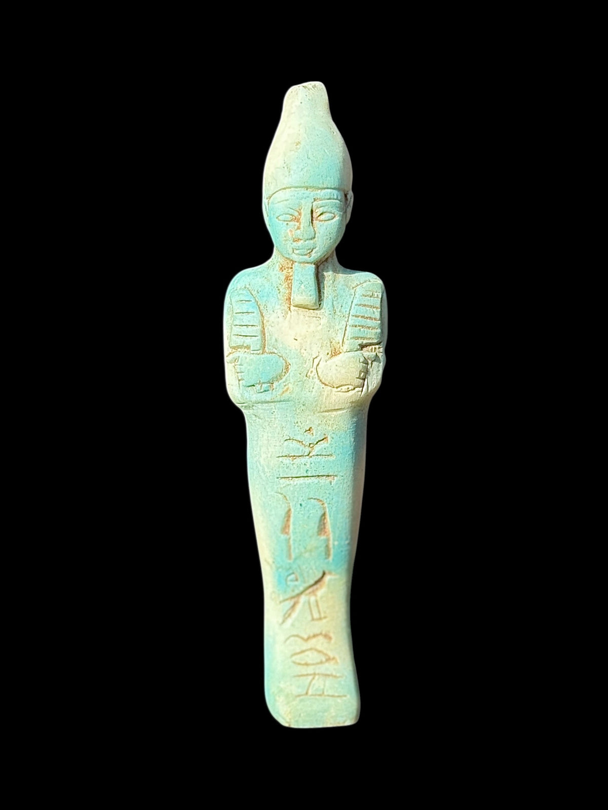 Front view of a pale turquoise and cream shabti with a tall crown, straight beard, crossed arms and incised markings against a black background.