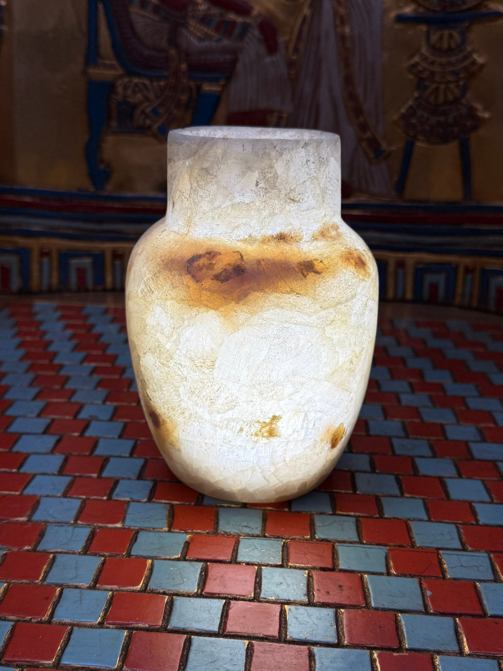 Small cream alabaster vase with a straight neck, rounded shoulders and a honey-coloured marking — illuminated view.