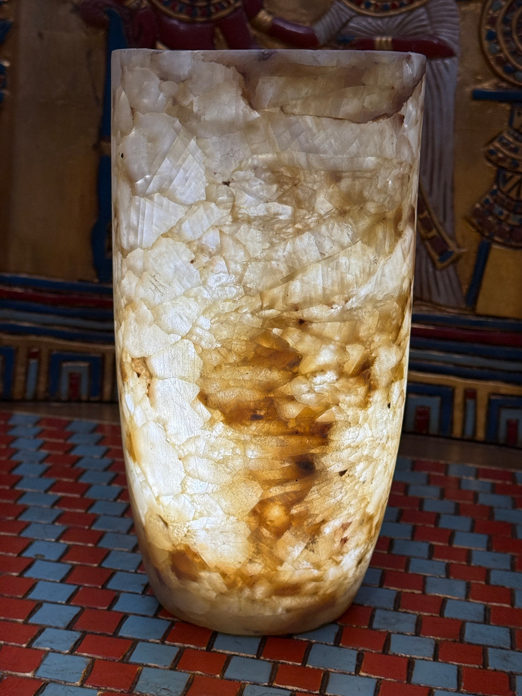 Slender cylindrical alabaster vase with layered cream, taupe and honey-coloured stone — illuminated view.