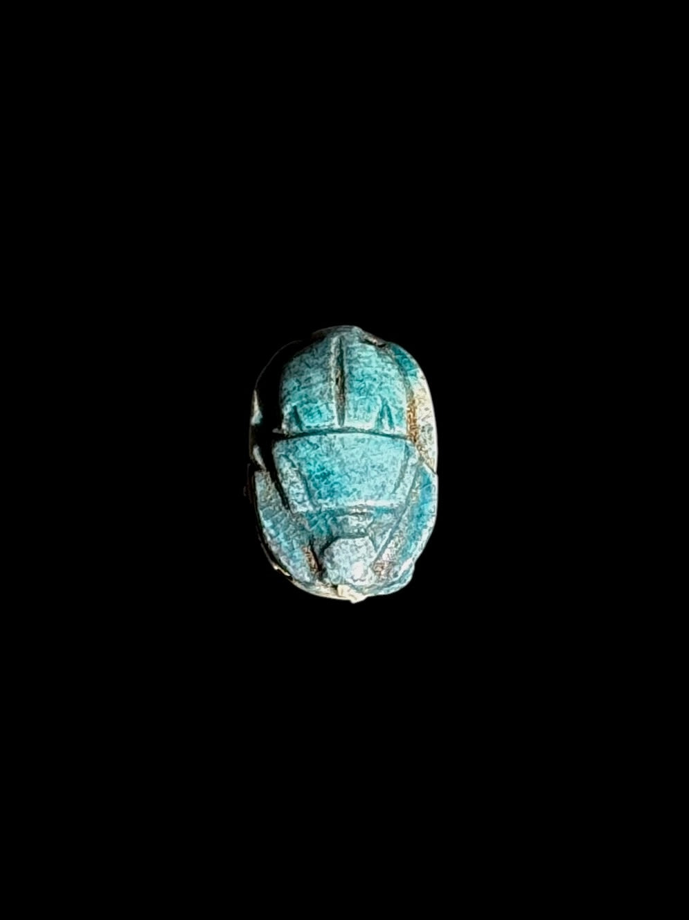 Turquoise-coloured beetle face of a stone scarab, showing outlined wing divisions and pale patches along the edges against a black background.