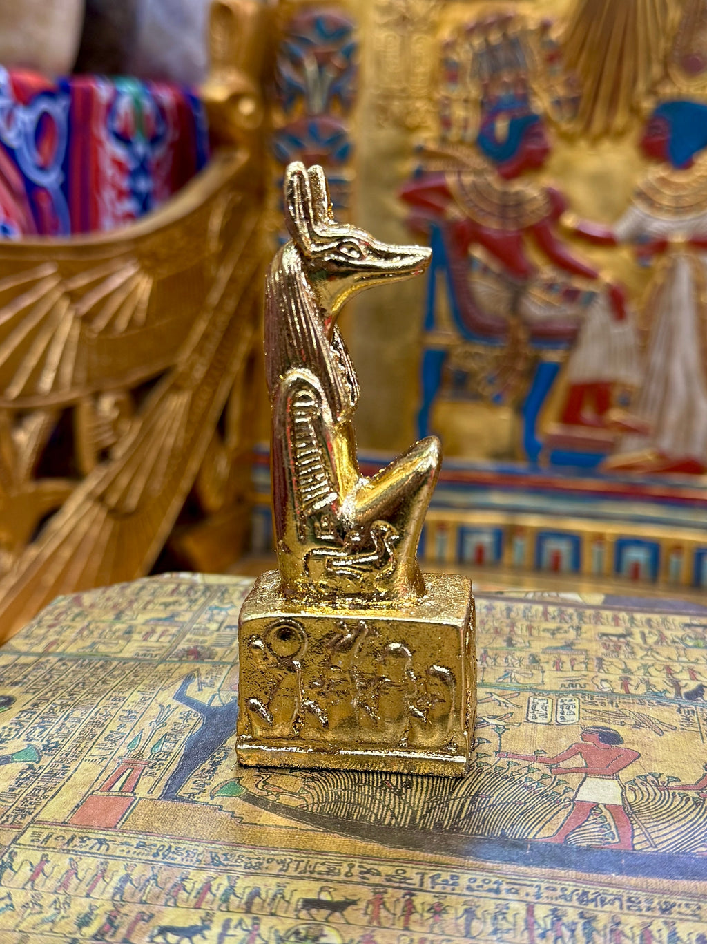 Side view of the gold finish Anubis kneeling statue with jackal head, upright ears and decorated square base.