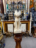 Front view of the cream Bastet cat statue on a wooden display pedestal, with other Egyptian statues in the background.