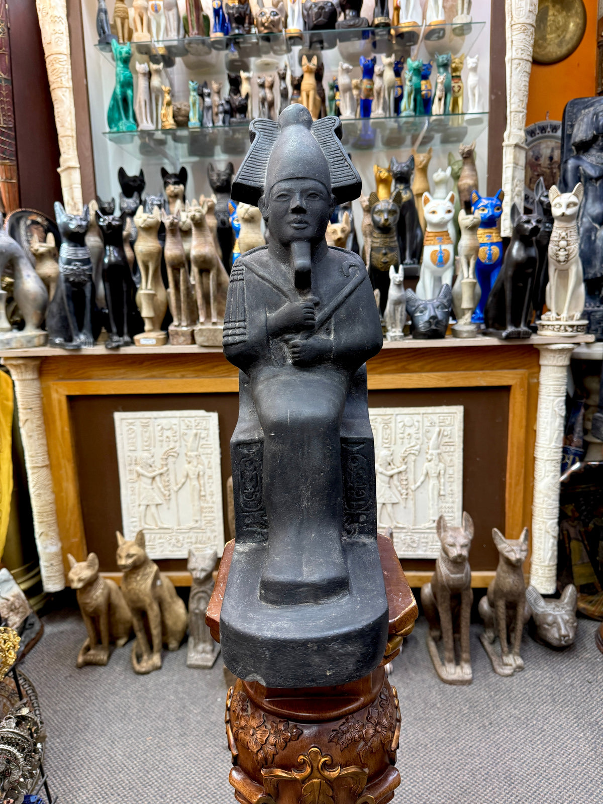 Front view of a dark seated Osiris statue with feathered crown, crossed arms, inscribed throne and extended base.