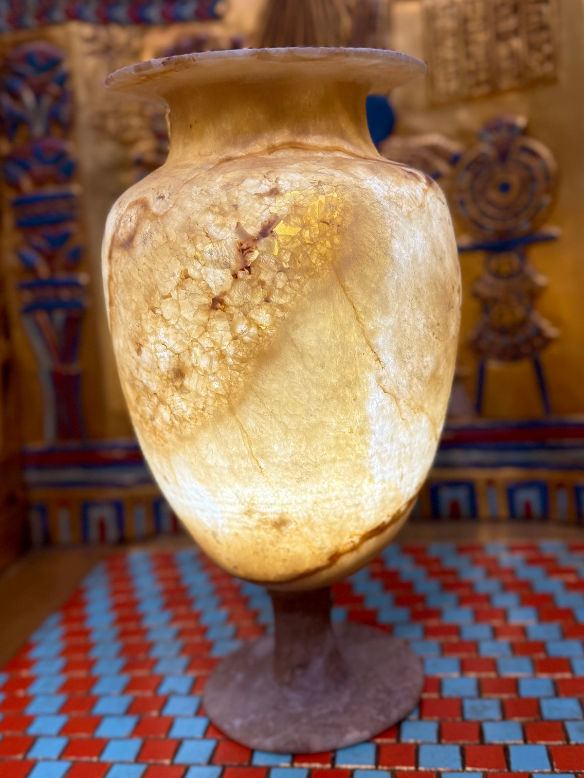 Cream alabaster pedestal vase with an oval body, slender stem, round foot and flared rim — illuminated view showing warm honey-coloured stone.