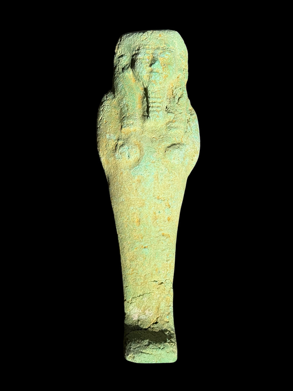 Front view of a pale green shabti with crossed arms, a long headdress, beard and rough, sandy-coloured patches against a black background.