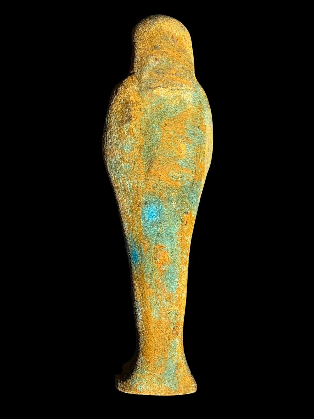 Back view of a shabti figurine showing a rounded head, tapering body and textured ochre surface with blue-green patches.