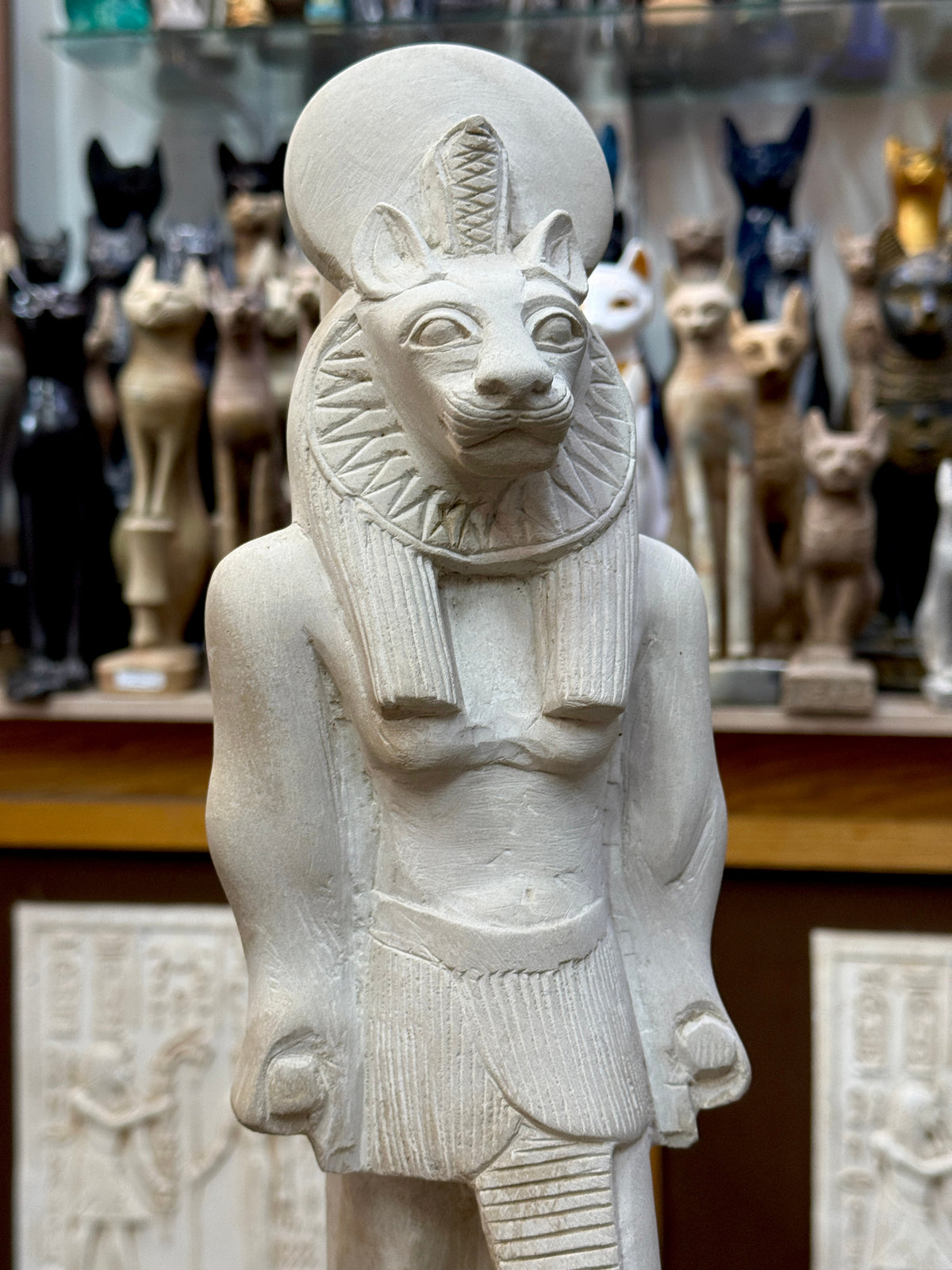 Close front view of Sekhmet's lioness face, cobra and sun disc, patterned collar, striated headdress and pleated garment.
