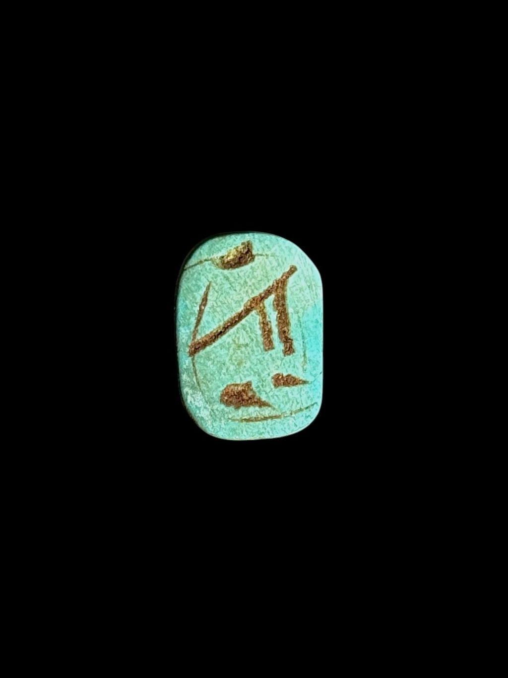 Underside of a blue-green stone scarab showing brown-toned incised markings within a rounded border against a black background.