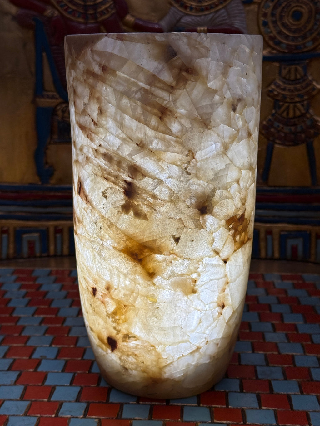 Slender cylindrical alabaster vase with layered cream, taupe and honey-coloured stone — illuminated view.
