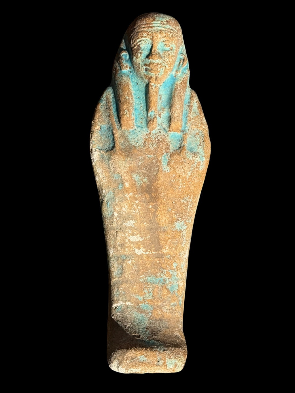 Front view of a stone shabti with a long headdress, elongated beard and turquoise patches across a sandy brown surface, against black.
