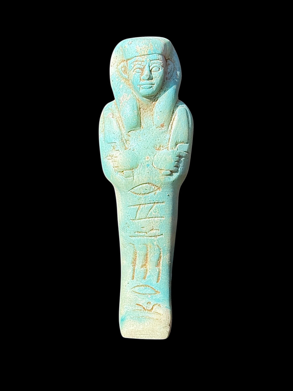 Front of turquoise-coloured stone shabti with a long headdress, crossed arms, incised markings and pale cream patches.