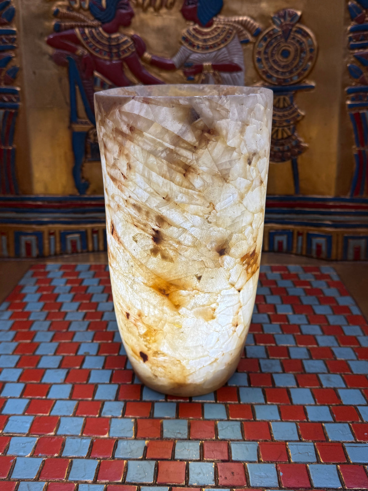Slender cylindrical alabaster vase with layered cream, taupe and honey-coloured stone — illuminated view.