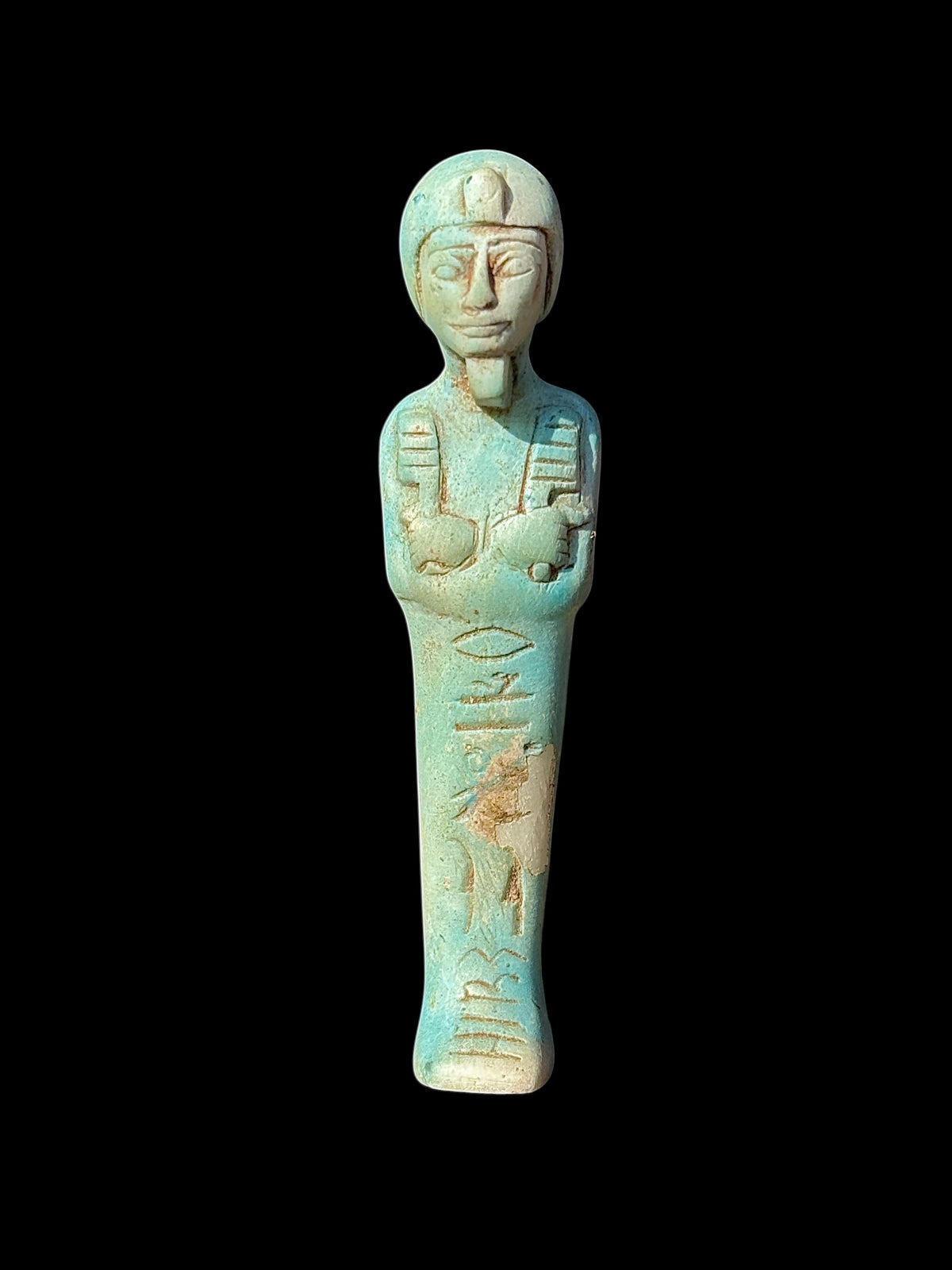 Front view of a turquoise-coloured shabti figurine with crossed arms, headdress, beard and incised markings, with cream and tan surface patches.