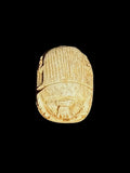 Top view of a pale cream and tan stone scarab with parallel grooves across its back and outlined beetle details against a black background.