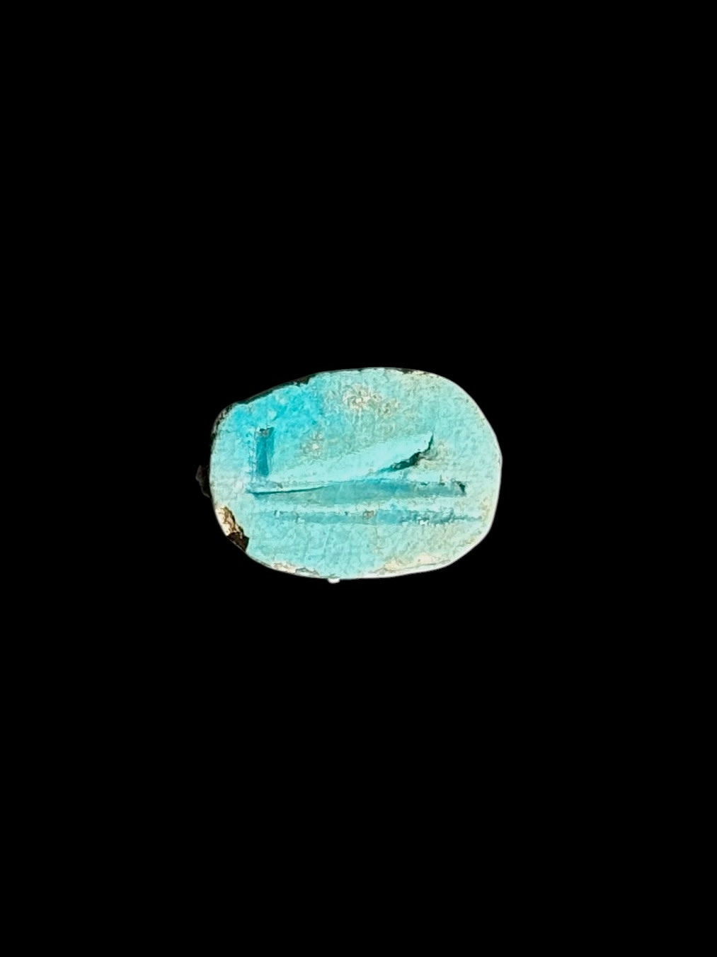 Underside of the stone scarab showing a turquoise-coloured oval surface with incised linear markings against a black background.