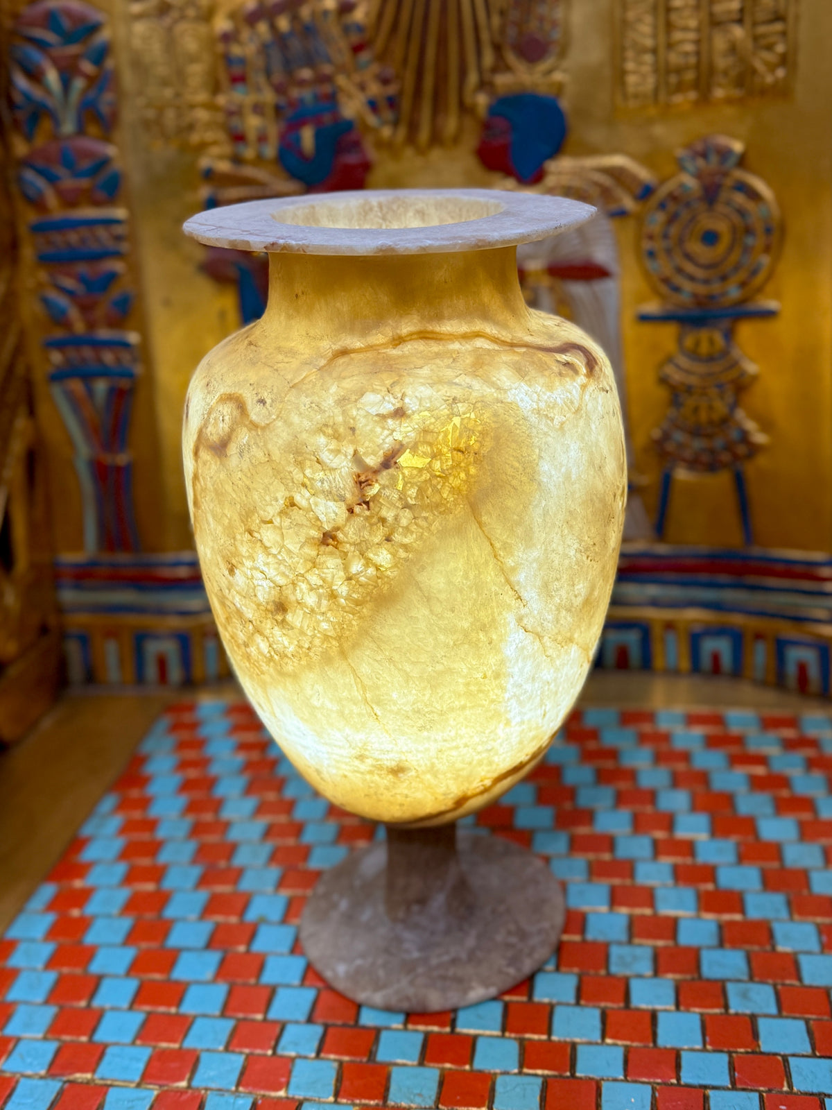 Cream alabaster pedestal vase with an oval body, slender stem, round foot and flared rim — illuminated view showing warm honey-coloured stone.