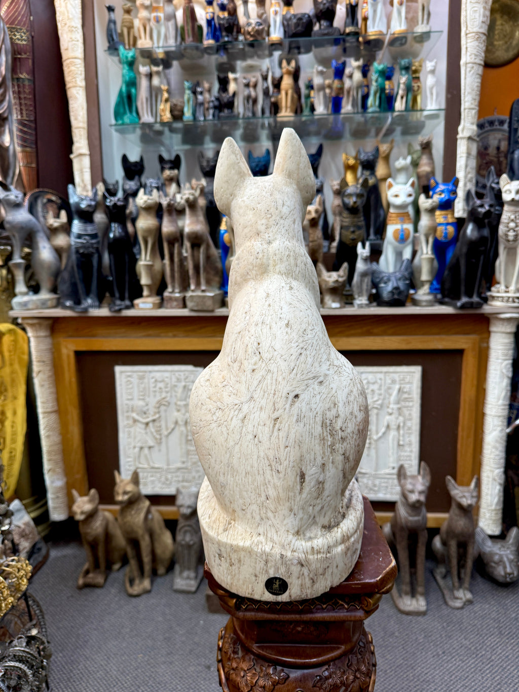 Rear view of the cream Bastet statue showing upright ears, rounded back, surface markings and a small label on the base.