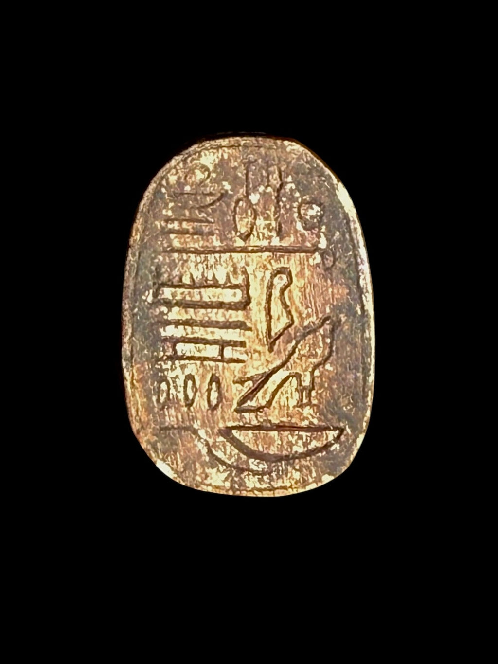 Oval underside of the stone scarab showing incised hieroglyphic markings, including a bird-shaped figure, on a gold-coloured surface.