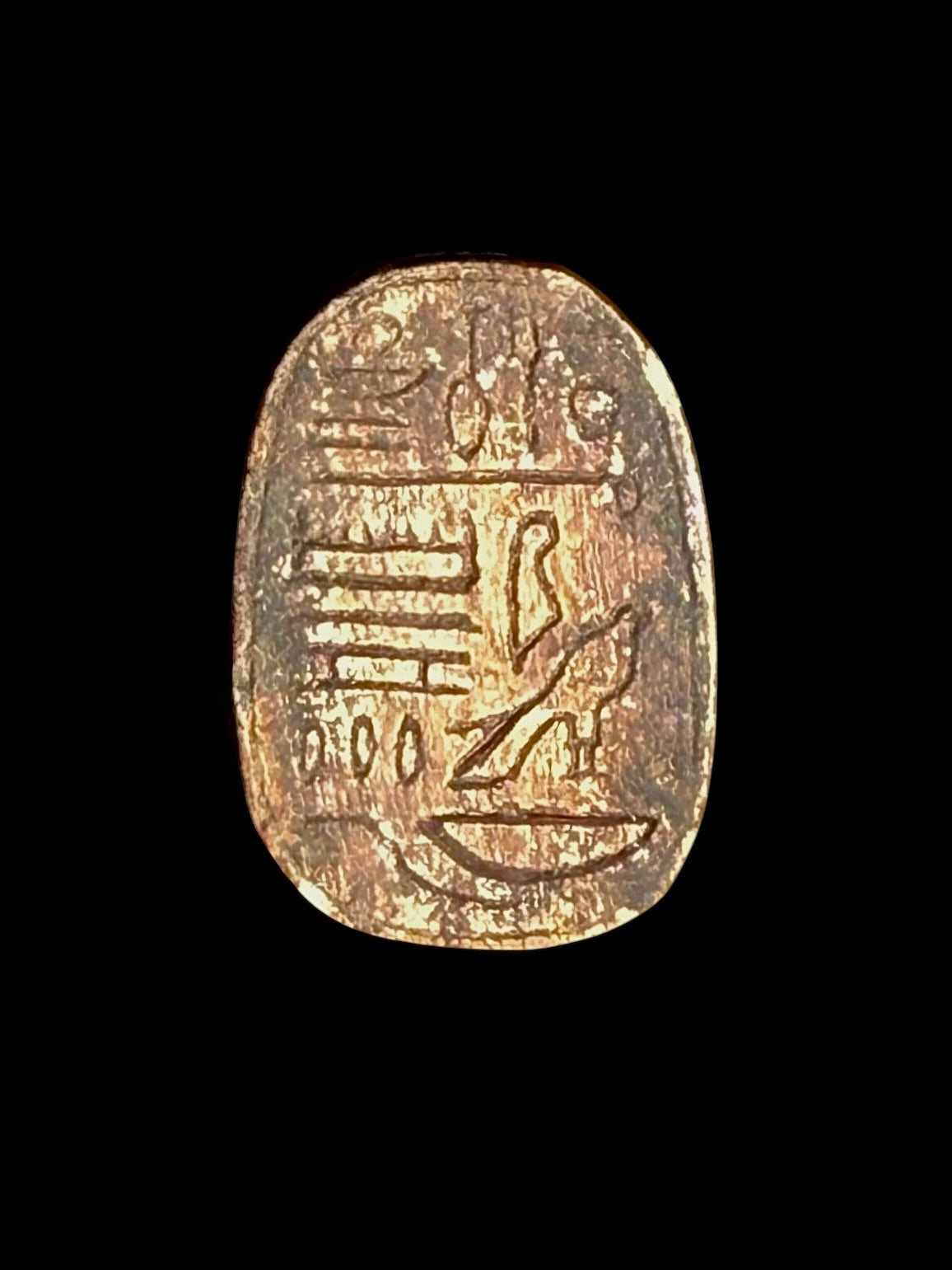 Oval underside of the stone scarab showing incised hieroglyphic markings, including a bird-shaped figure, on a gold-coloured surface.