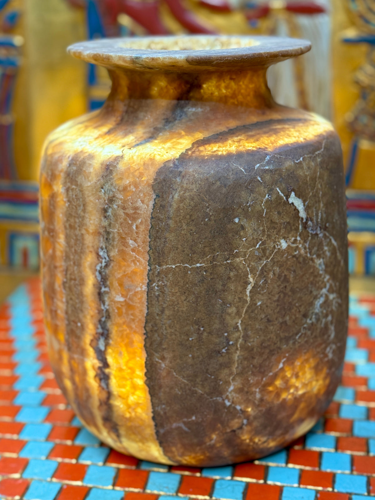 Squared alabaster vase with rounded corners, smoky brown stone, lighter vertical stripes and a flared rim — illuminated view showing amber-coloured stone.