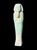 Front view of a pale turquoise and cream shabti figurine with a headdress, long beard, crossed arms and recessed markings down the body.