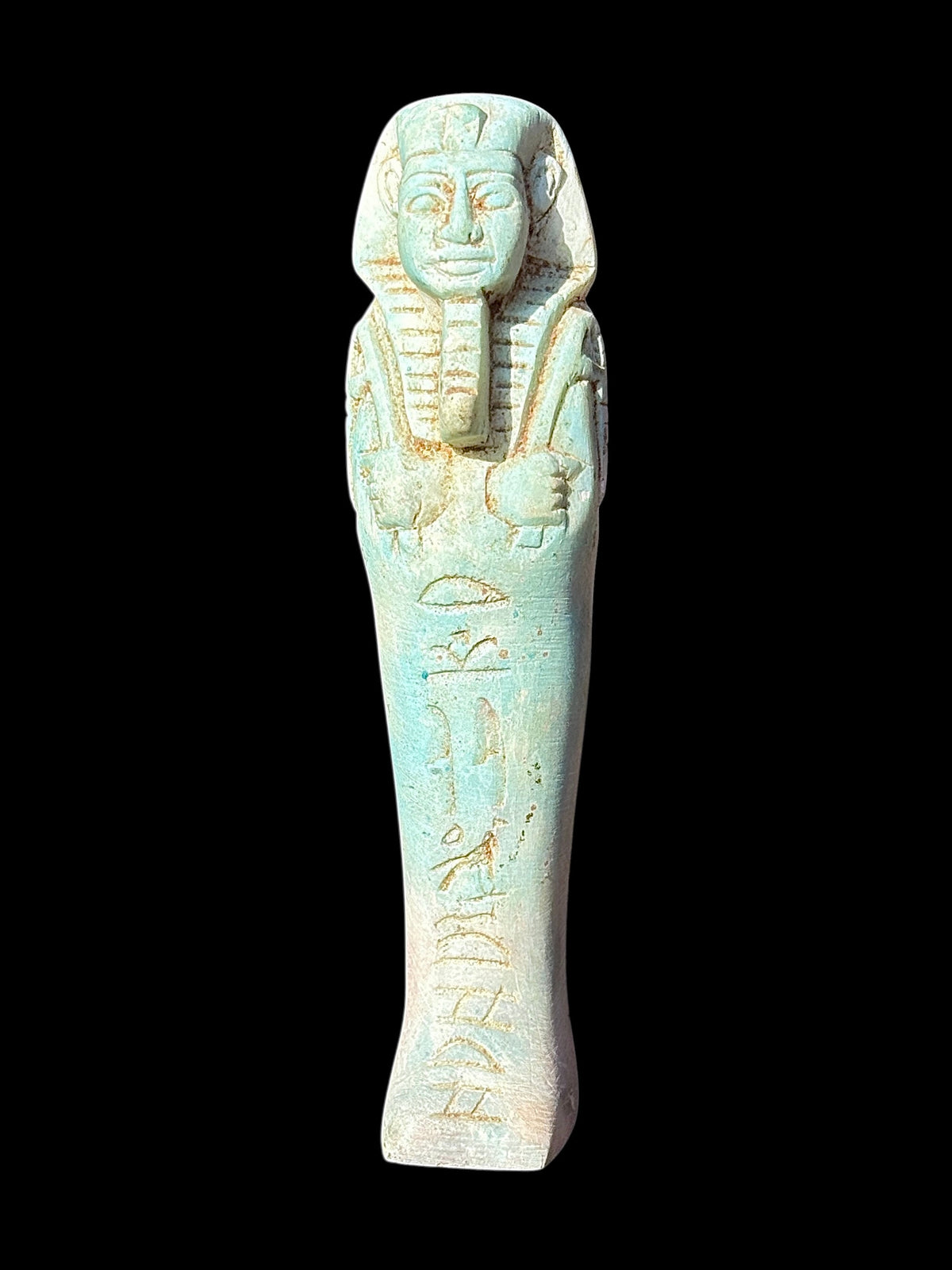 Front view of a pale turquoise and cream shabti figurine with a headdress, long beard, crossed arms and recessed markings down the body.