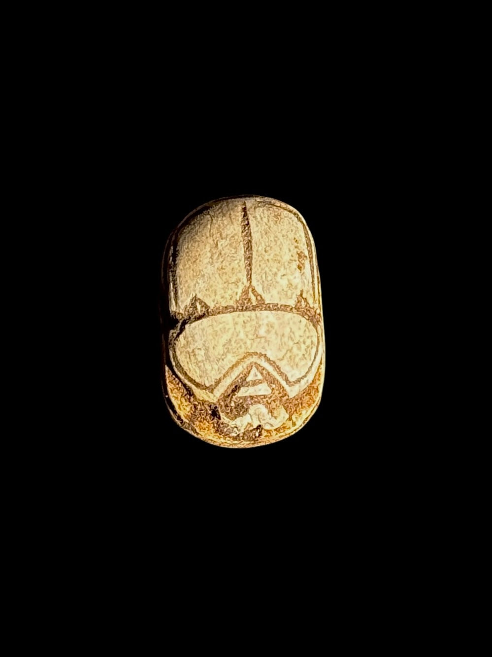 Top view of a cream-coloured stone scarab with brown incised lines defining the beetle’s back against a black background.