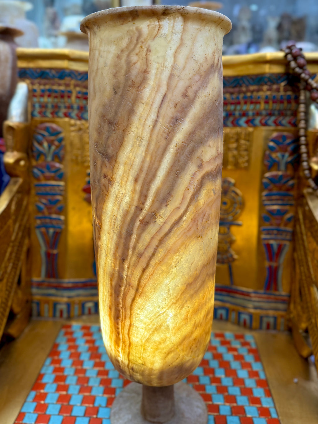 Tall cylindrical alabaster pedestal vase with flowing cream and honey-coloured ribbons, short stem and round foot — illuminated view.