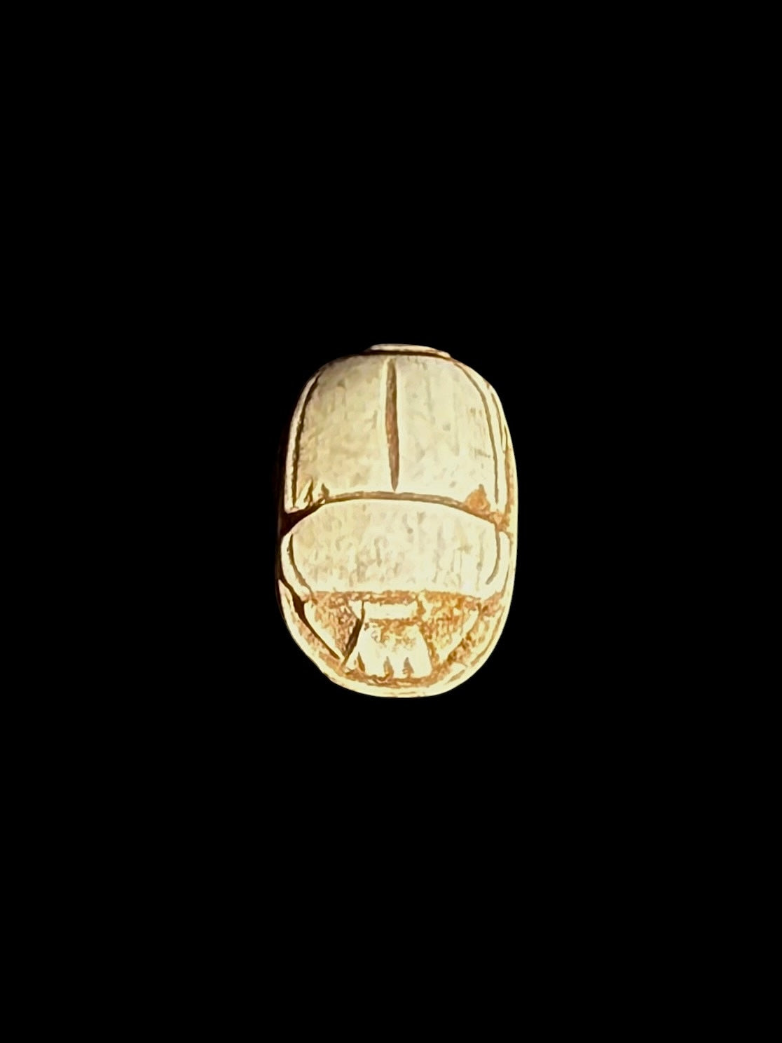 Top view of a cream-coloured stone scarab with ochre-brown lines defining the wings and beetle body against a black background.