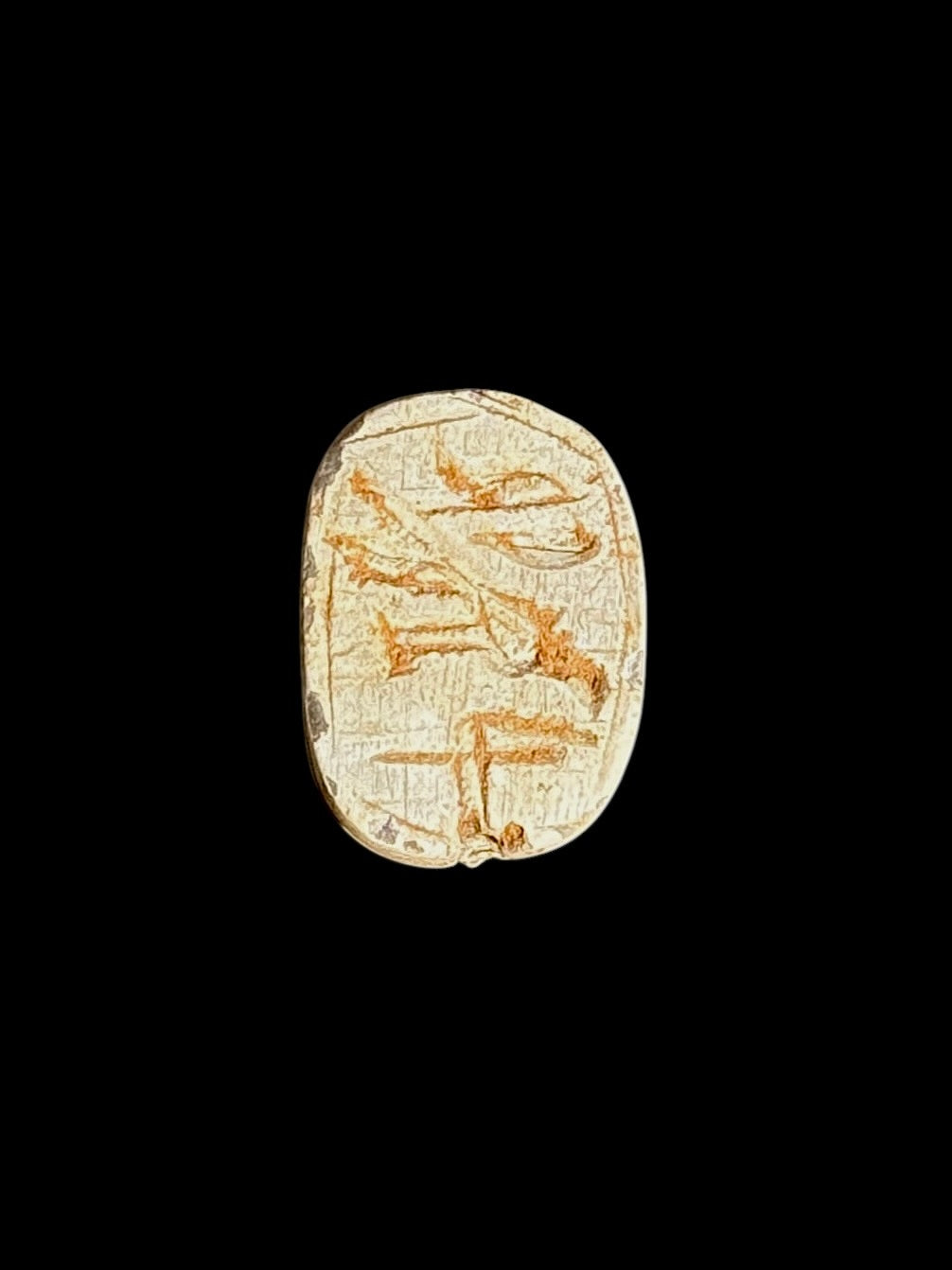 Underside of the stone scarab showing ochre-brown incised Egyptian-style markings within an oval border against a black background.