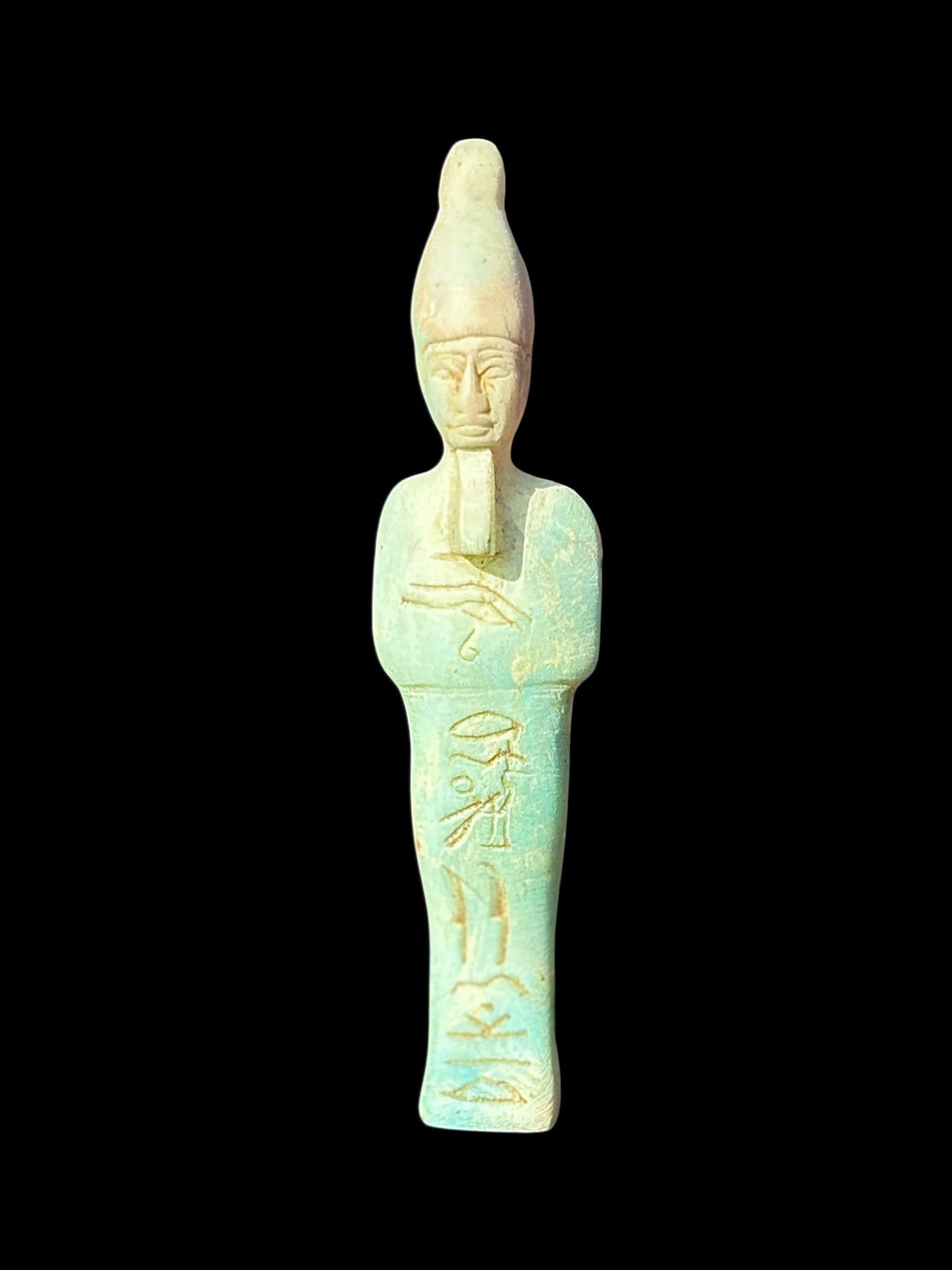 Front view of a pale turquoise-green and cream shabti with a tall crown, long beard, crossed arms and incised markings down the body.
